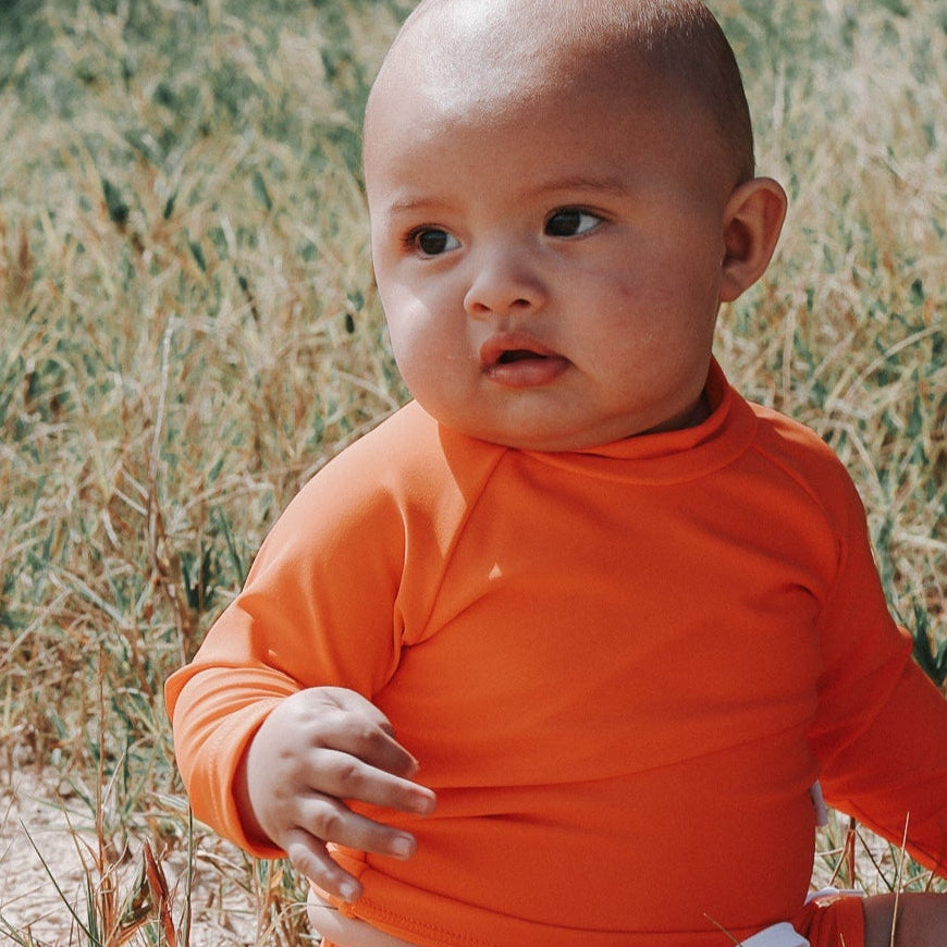 Baby in an bright orange swim shirt and swim short sitting in a sandy field