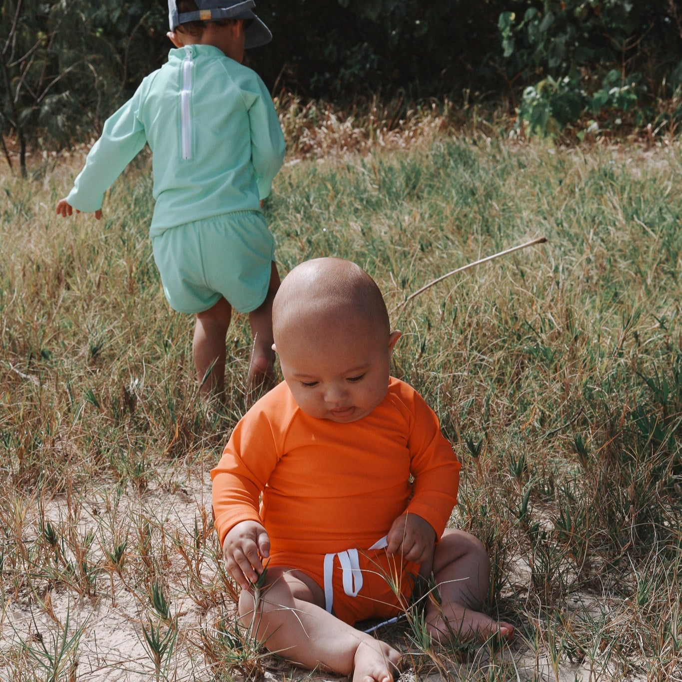 Two children in outdoor clothing walking through a grassy area with trees in the background.