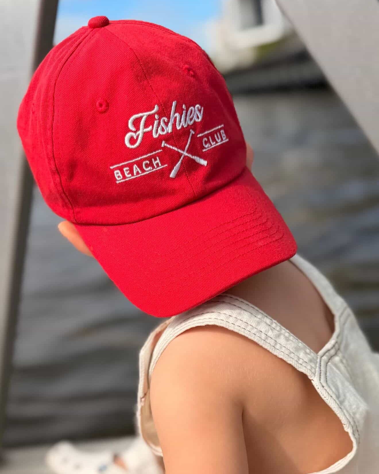 Child wearing a red cap with 'Fishies Beach' text, sitting by water.