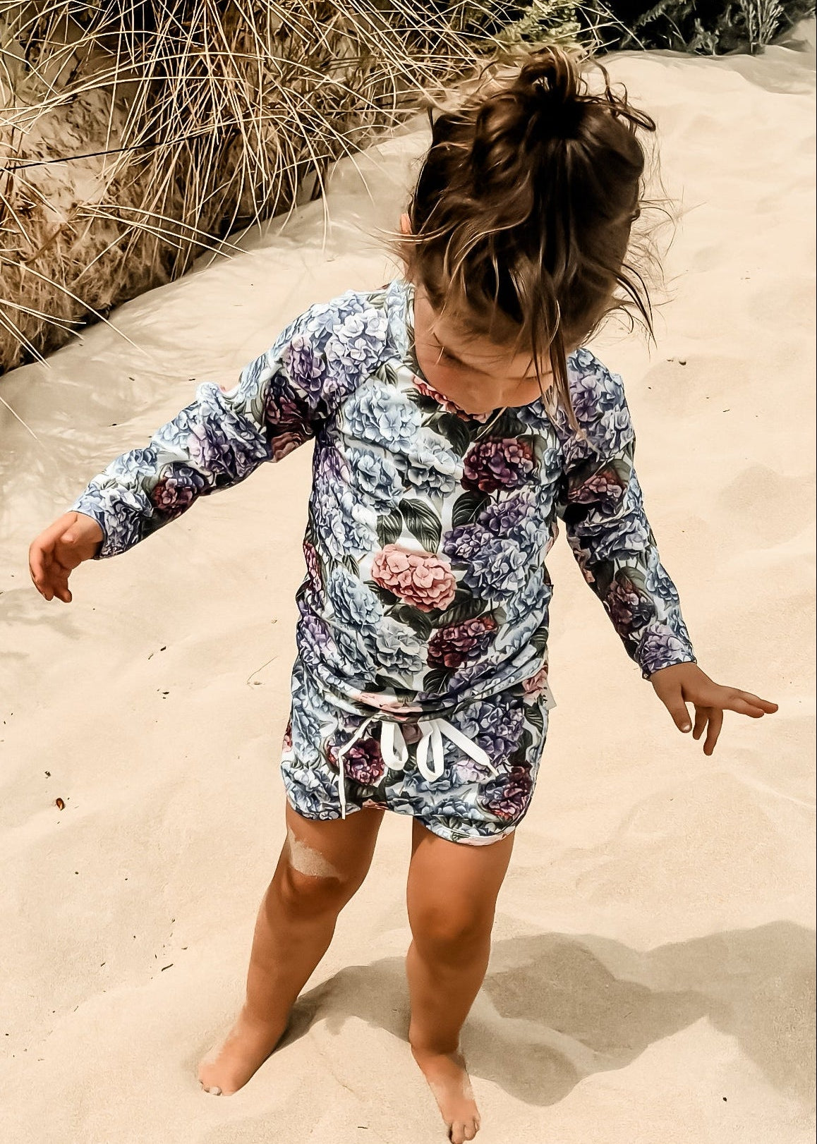 Child in a floral girls swimsuit standing on sand with plants in the background