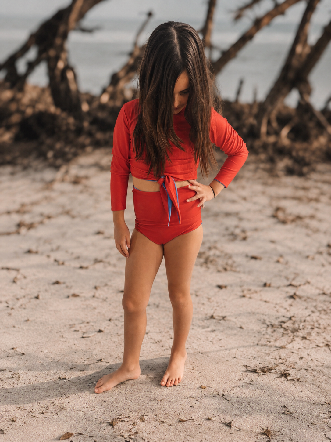 Young girl in a red swimsuit standing on a sandy beach with driftwood in the background