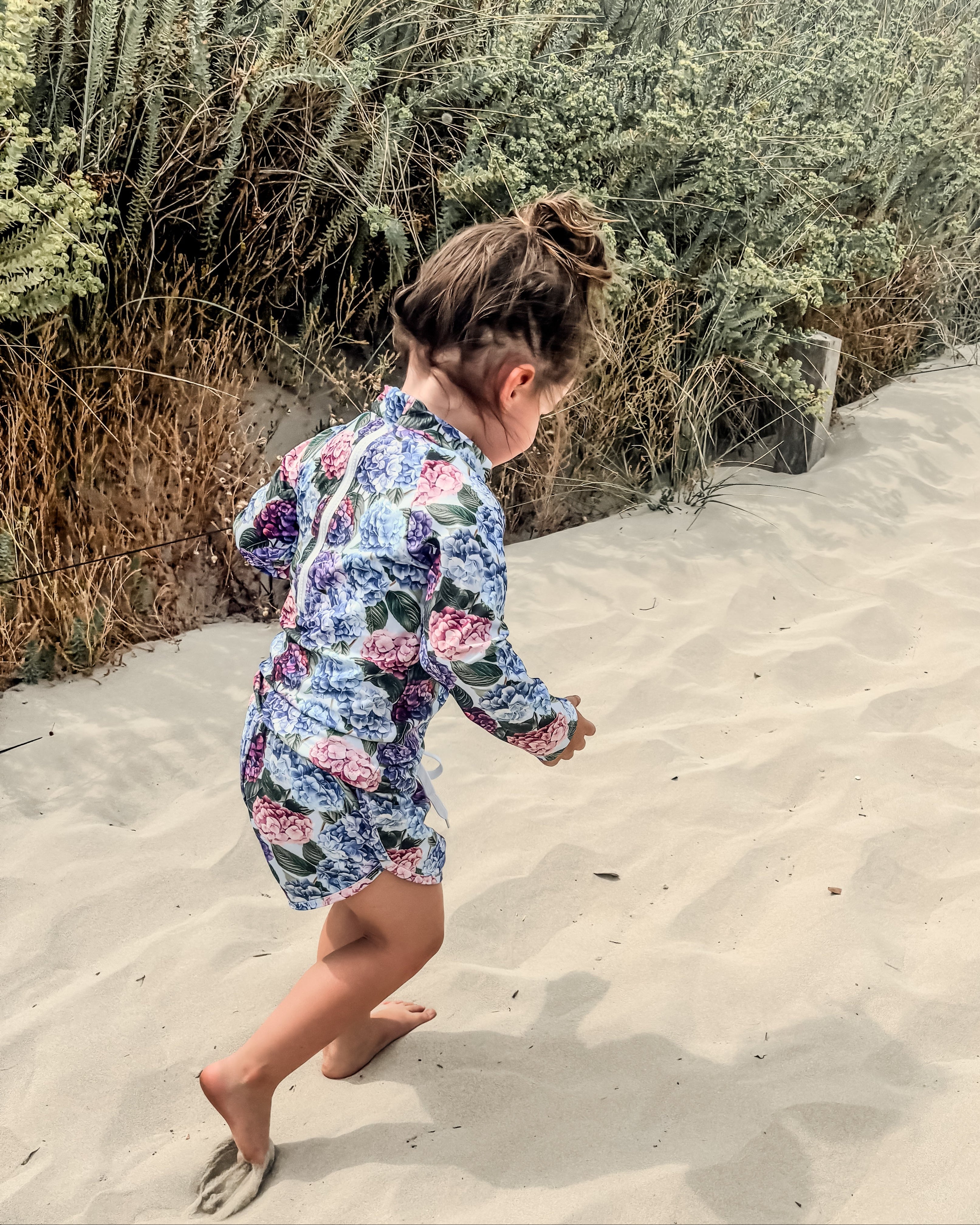 toddler girl in a floral rash set walking on sand with plants in the background