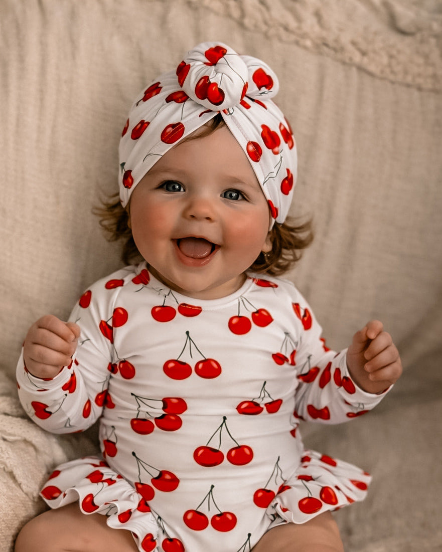Baby wearing a cherry-patterned swimsuit outfit and headband sitting on a beige surface.
