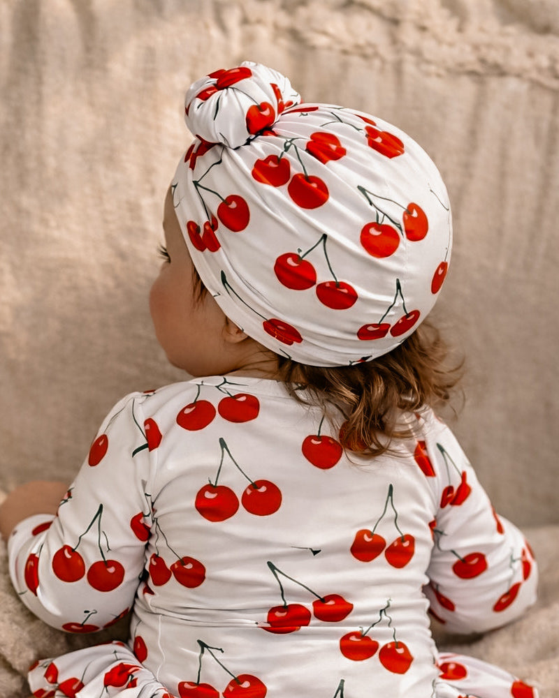 Baby in a cherry-themed long sleeved swim suit baby bathers with matching headband sitting on a beige couch.