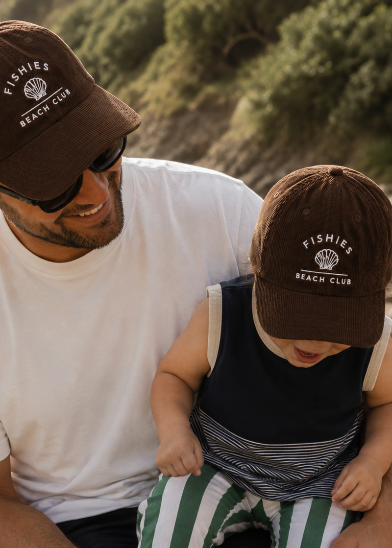 A man and a child sitting together on a wooden bench at the beach, both wearing matching brown baseball caps with white embroidered text that reads 'FISHIES BEACH CLUB'.
