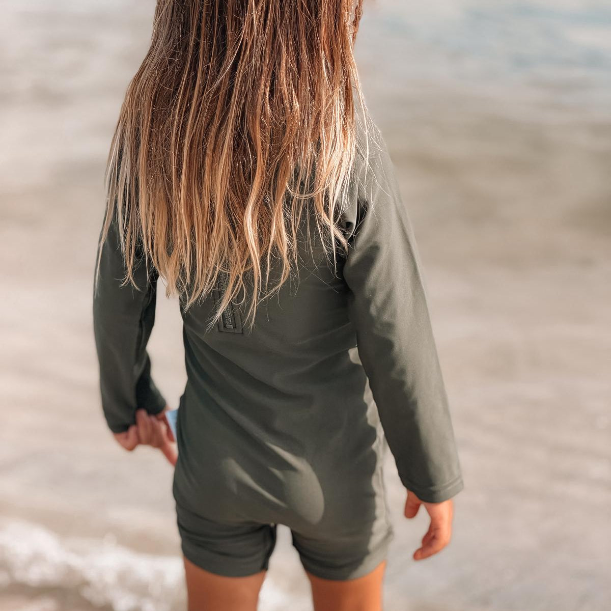 Child wearing a green long-sleeve rash guard and shorts standing on a beach.