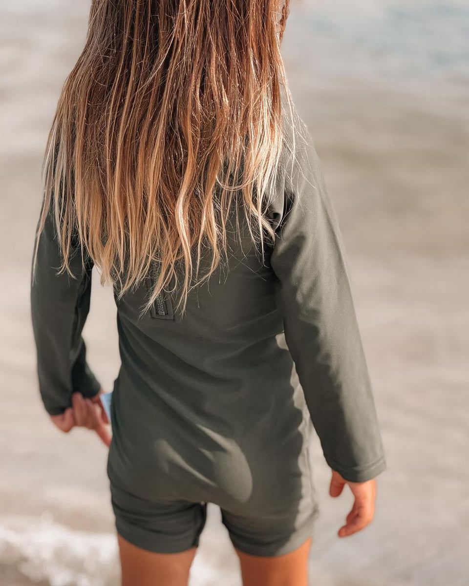 Child wearing a green long-sleeve rash guard and shorts standing on a beach.