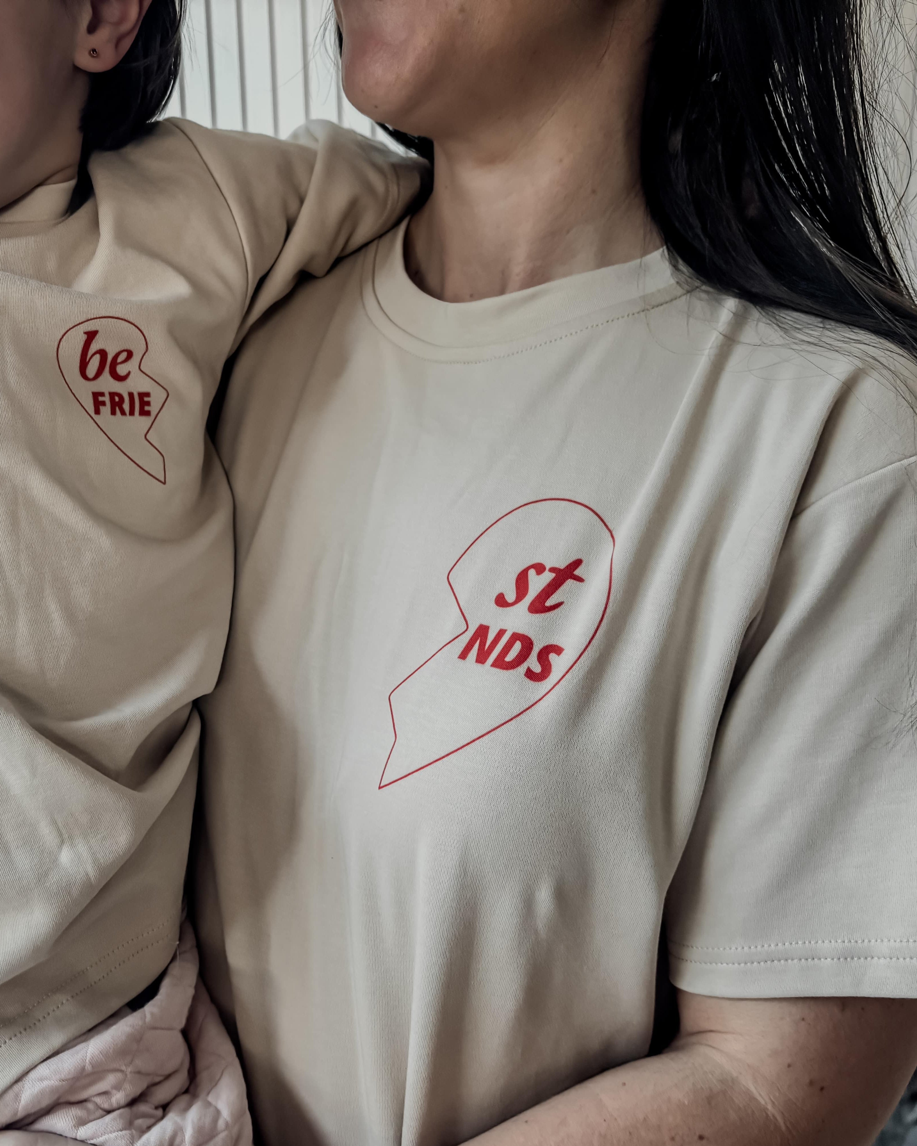 Two people wearing matching beige t-shirts with red heart designs and text.