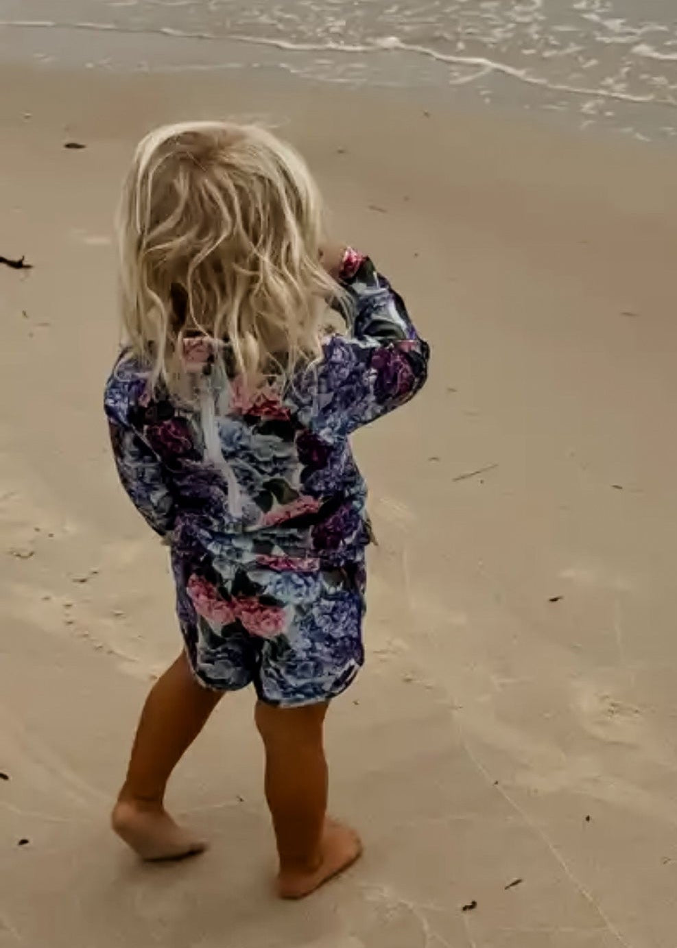 Child in a floral girls rash set outfit standing on a sandy beach with waves in the background