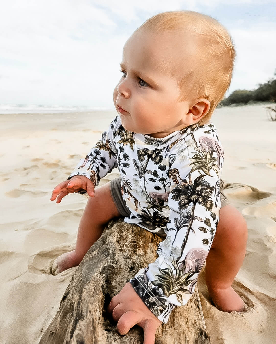 Baby sitting on a log at the beach wearing a boys bright rash guard set.