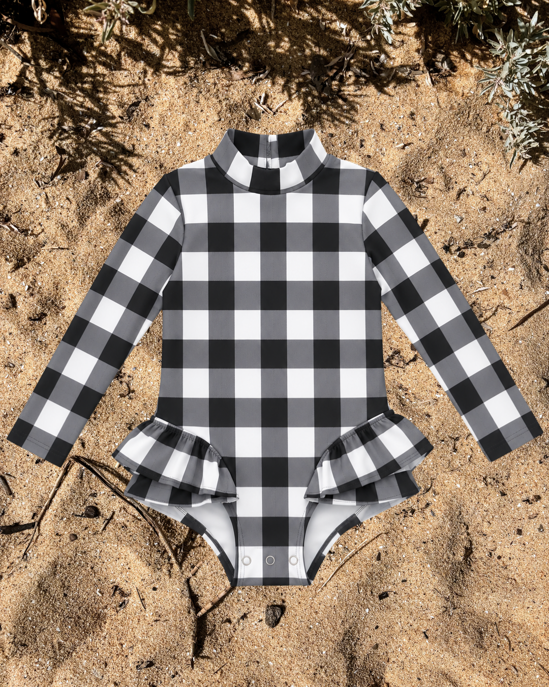 A long-sleeve baby and girls swimsuit with a black and white check gingham pattern, featuring a mock neck and ruffled leg hems, laid flat on sandy ground.