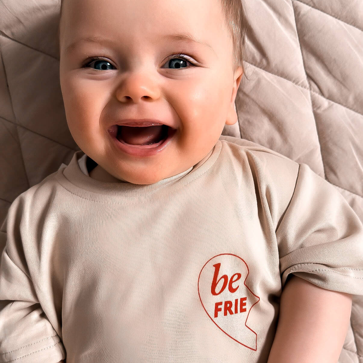 Baby wearing a shirt with 'be FRIE' text on a quilted surface