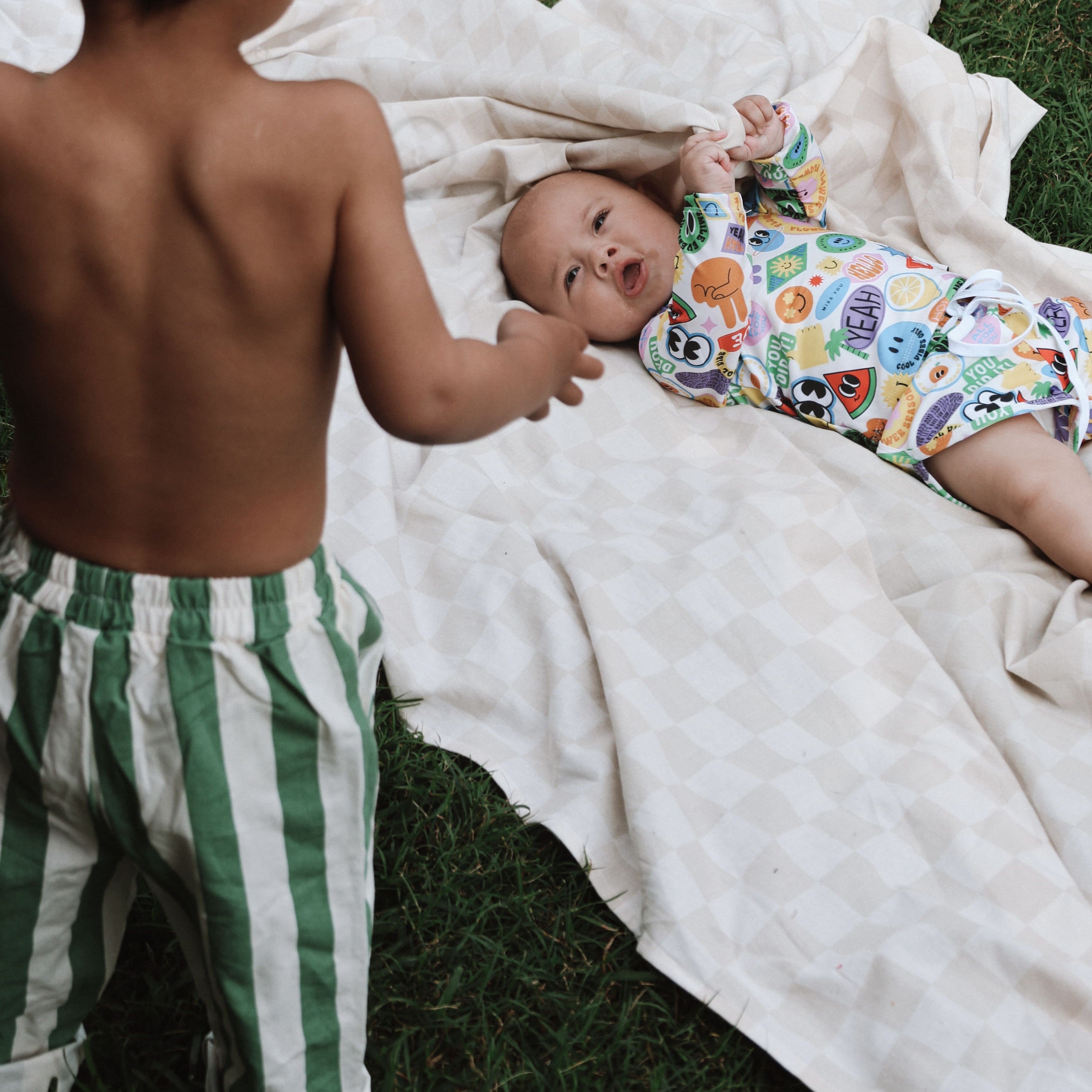 Two children on a blanket outdoors, one child is lying down and the other is standing.