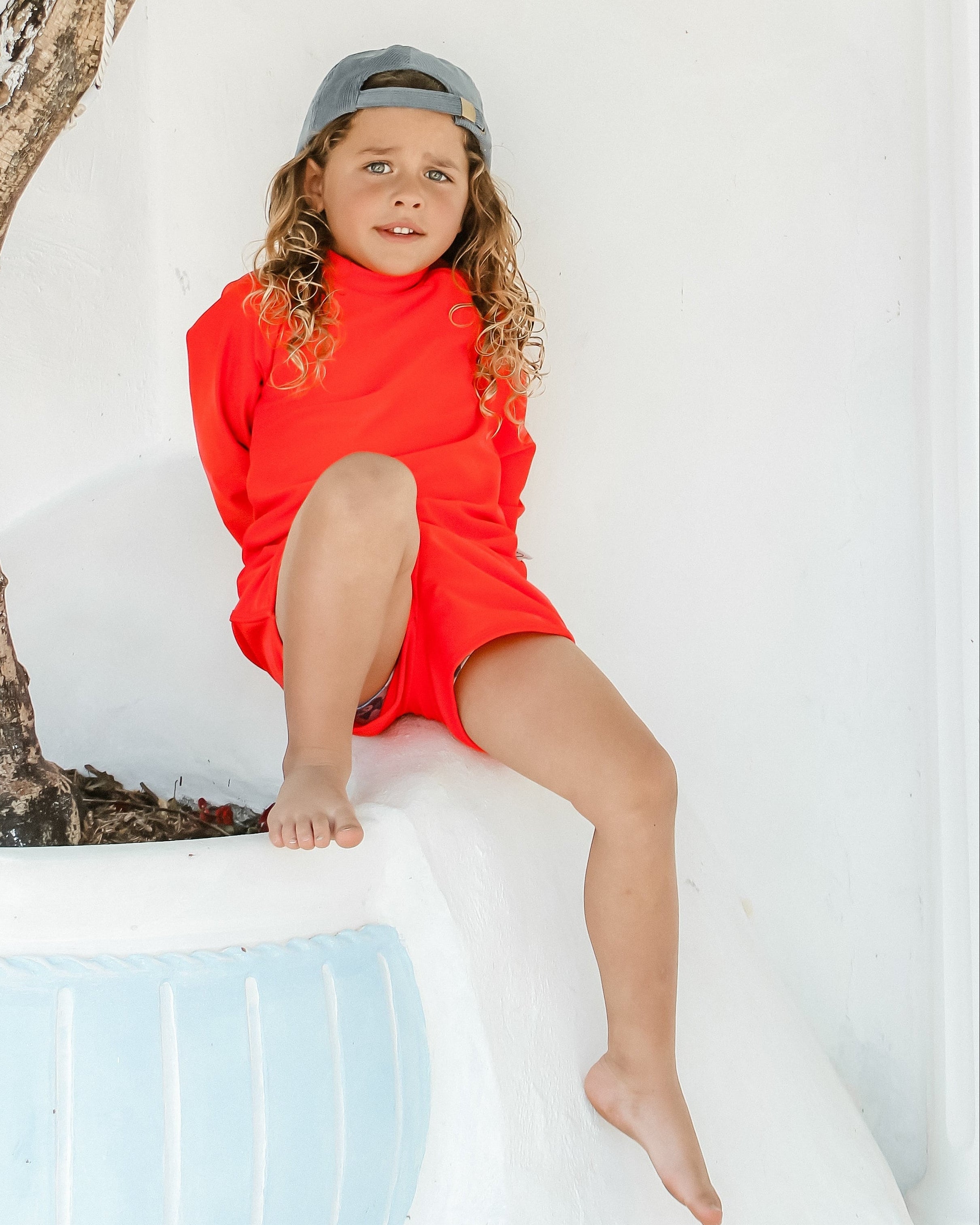 Child in a bright hi vis red rash suit swim set sitting on a white ledge with a tree and white wall in the background