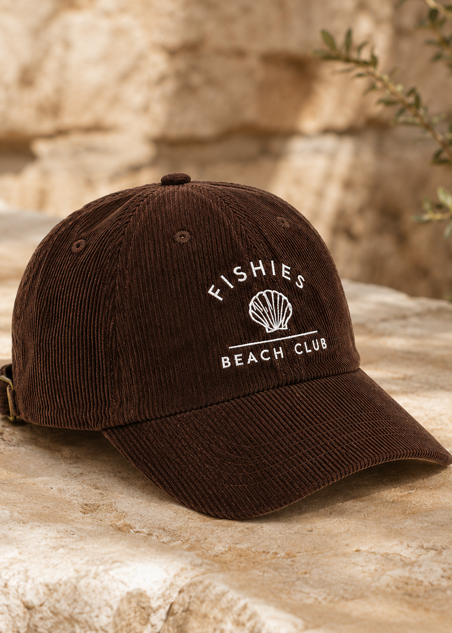 A dark brown corduroy baseball cap with white embroidered text reading 'FISHIES BEACH CLUB' and a shell graphic, resting on a light stone surface outdoors.