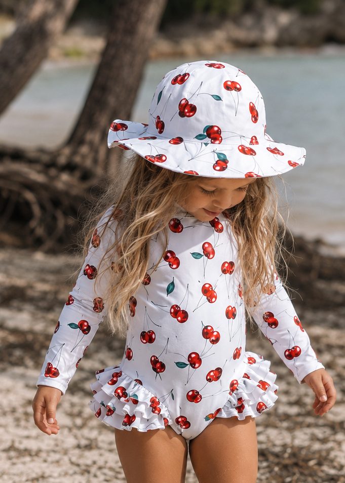A young girl stands on a sandy beach wearing a white long-sleeve one-piece swimsuit and matching sun hat, both printed with a red cherry pattern. Trees and water are visible in the background.