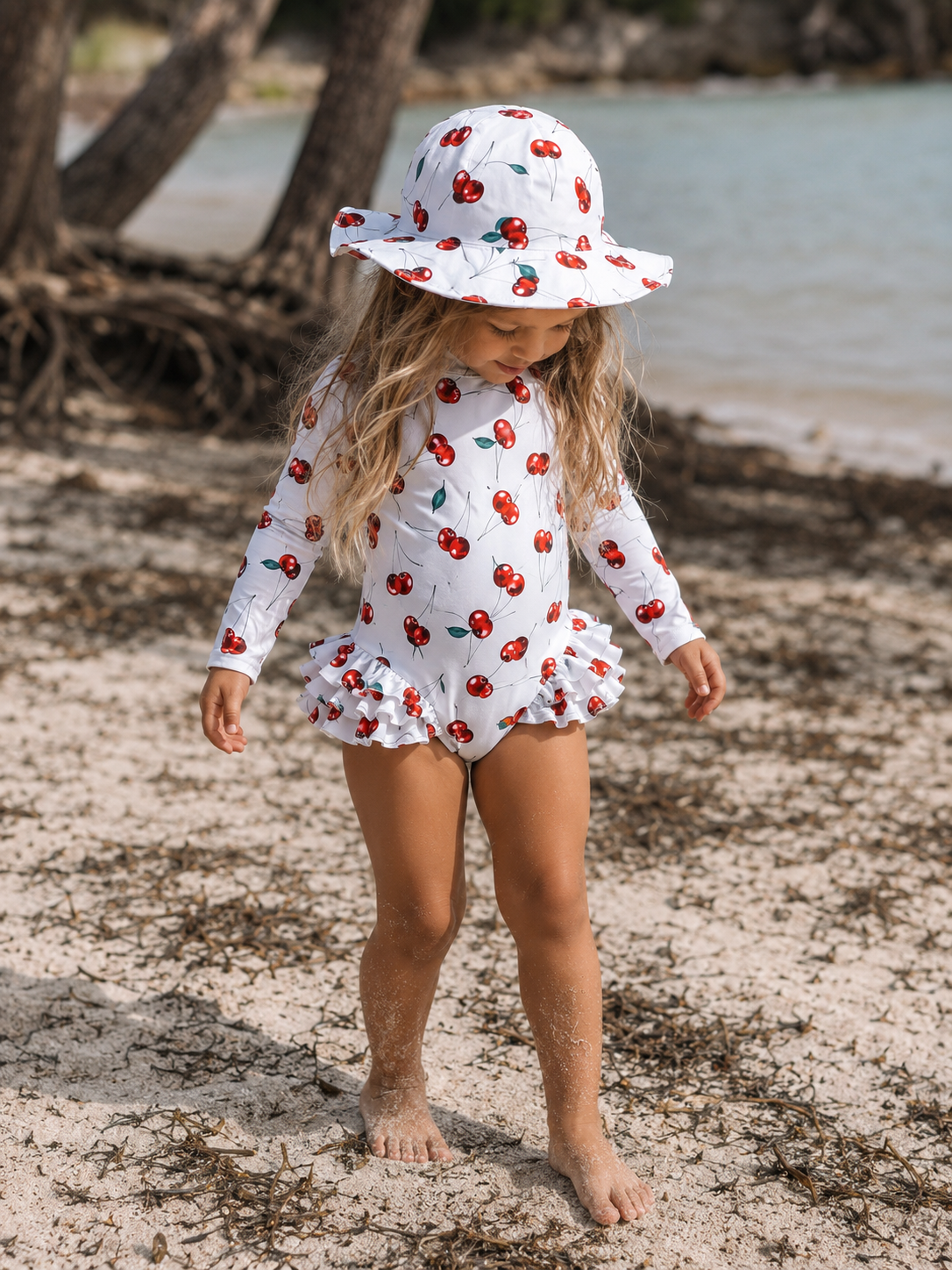 A young girl stands on a sandy beach wearing a white long-sleeve one-piece swimsuit and matching sun hat, both printed with a red cherry pattern. Trees and water are visible in the background.