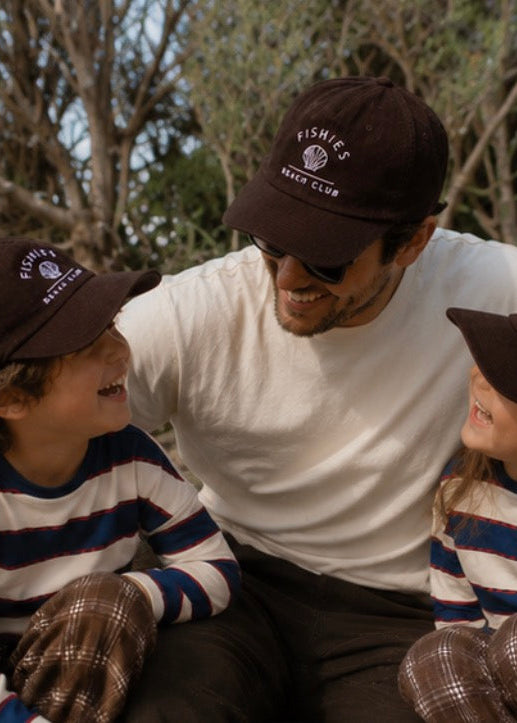 A man and two children wearing matching dark brown baseball caps with 'FISHING CLUB' printed in white on the front, posed outdoors among trees. The man wears a white t-shirt and dark pants, while the children wear striped long-sleeve shirts and plaid pants.
