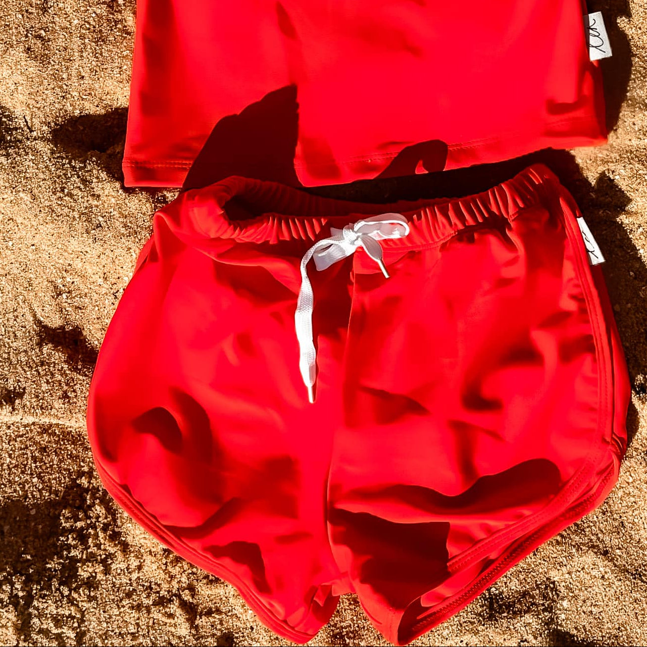 Bright Red children's rash set swimsuit set on a sandy surface with plants in the background
