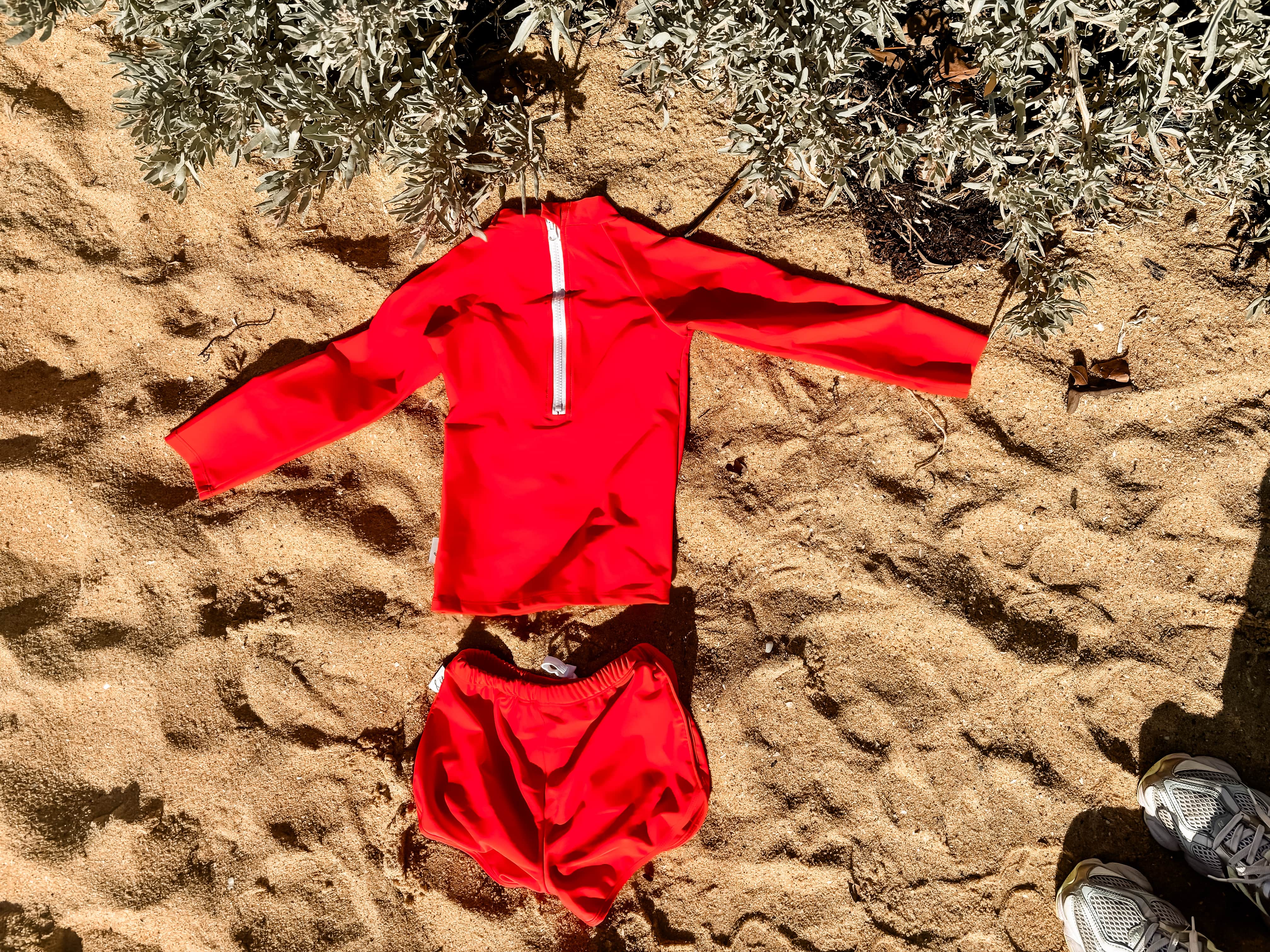 bright boys rash set Red outfit laid out on sandy ground with plants in the background
