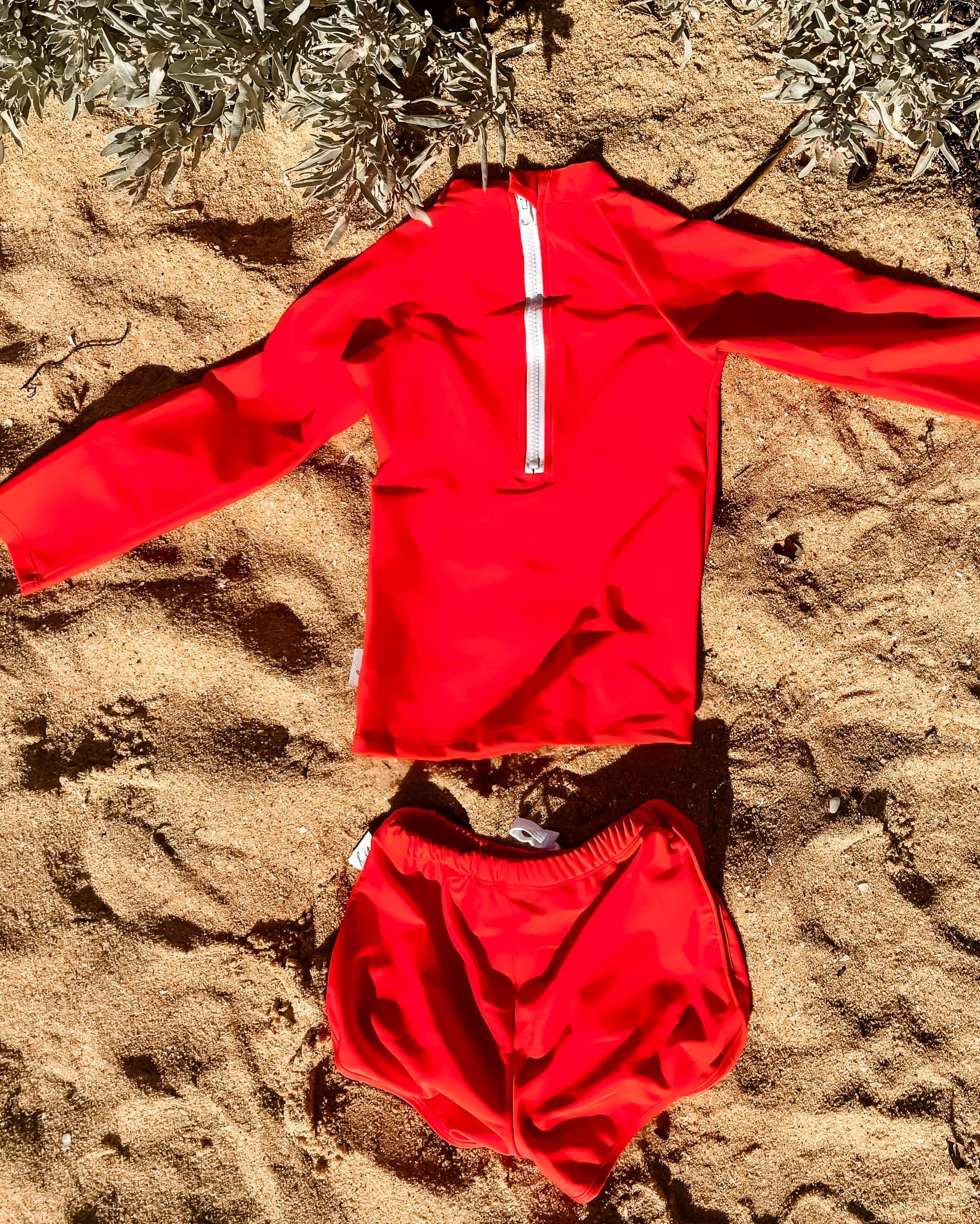 bright swimsuit for boys in a Red outfit laid out on sandy ground with plants in the background