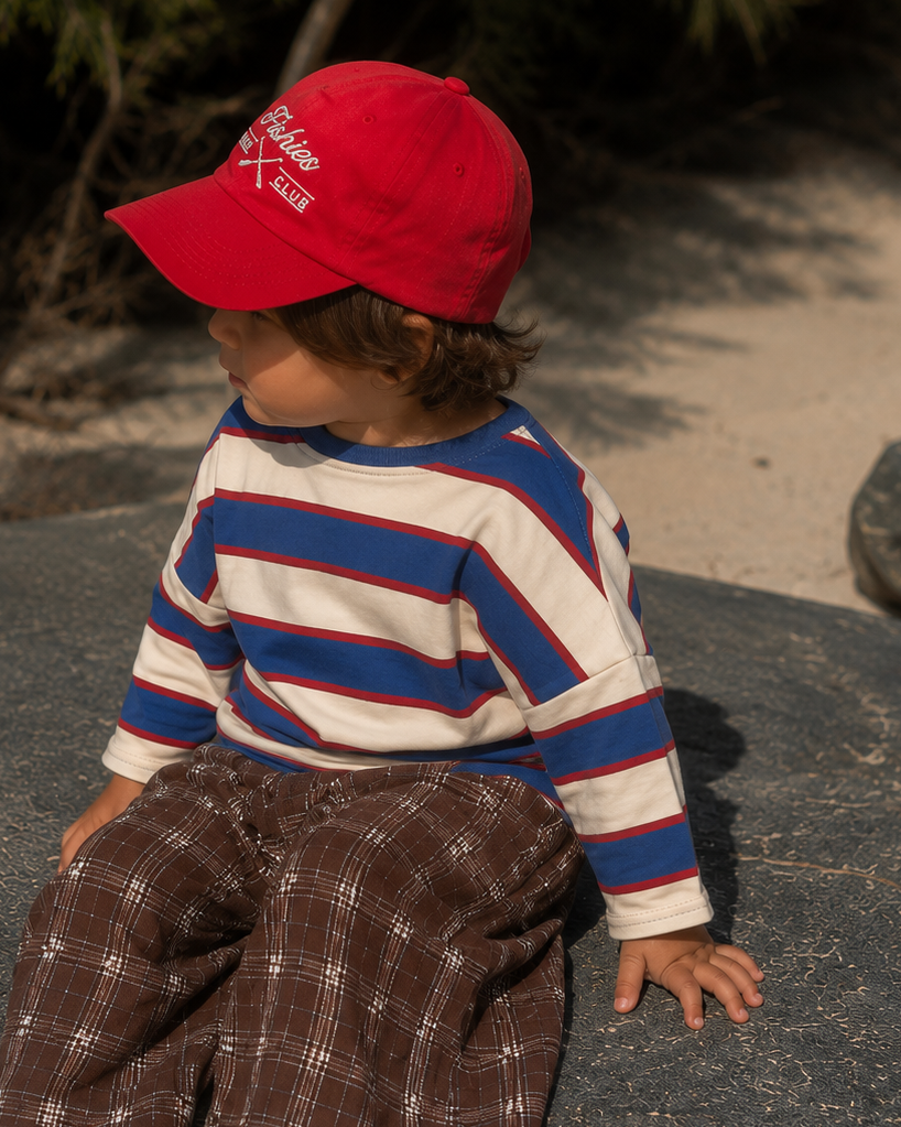 A toddler sitting outdoors on a large dark stone, wearing a bright red baseball cap with white embroidered text, a cream long-sleeve shirt with blue and red horizontal stripes, and brown plaid pants.
