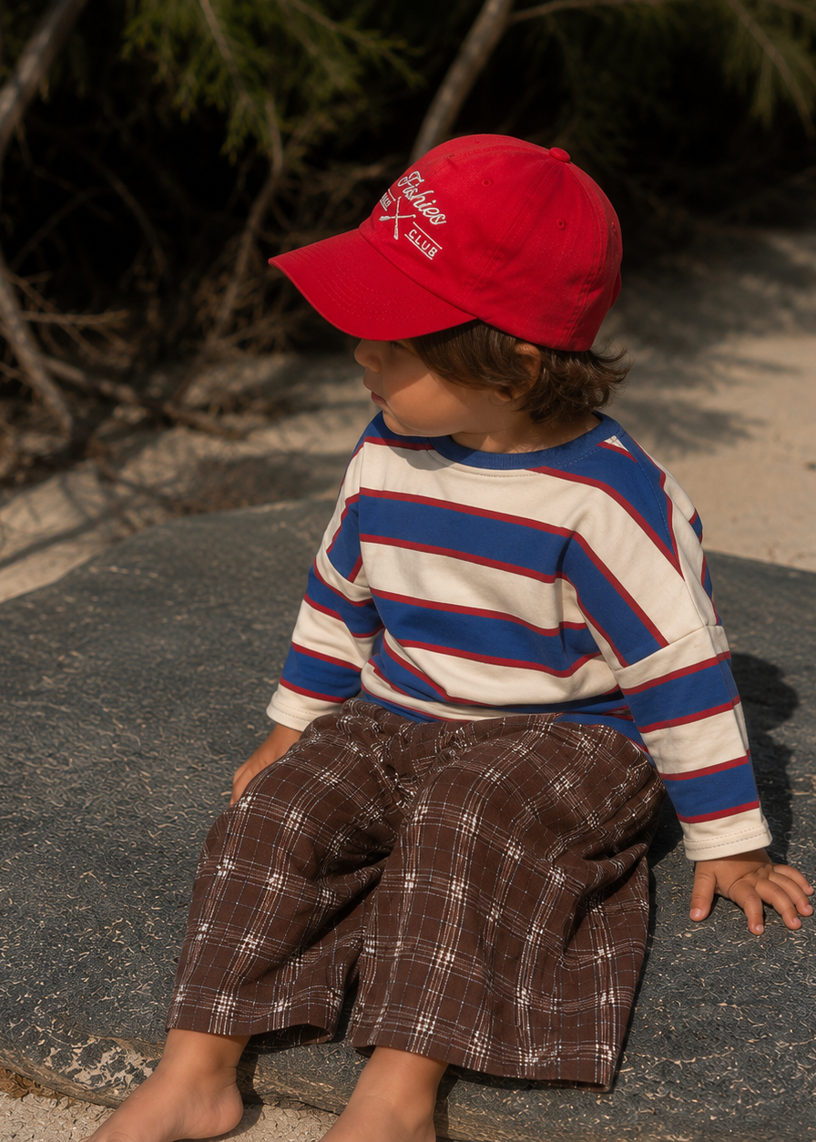 A toddler sitting outdoors on a large dark stone, wearing a bright red baseball cap with white embroidered text, a cream long-sleeve shirt with blue and red horizontal stripes, and brown plaid pants.