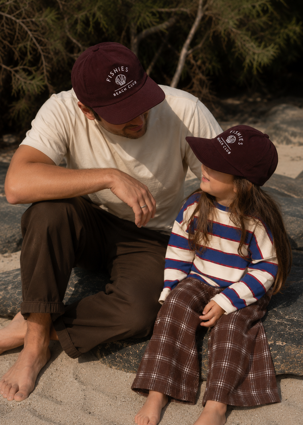 A man and a young girl sitting on rocks outdoors, both wearing matching dark maroon baseball caps with white embroidered 'FISHIES APPAREL CO' logos. The man wears a cream t-shirt and brown pants, while the girl wears a cream, blue, and brown striped long-sleeve top and brown plaid pants.