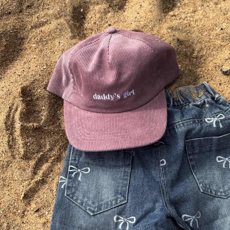Pink cap with 'daddy's girl' text on sand next to blue jeans with bow details