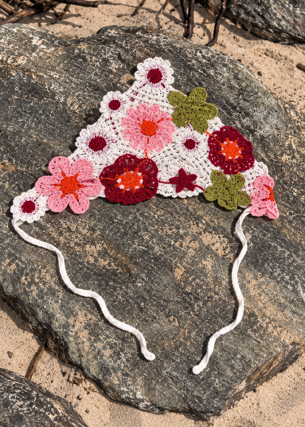 A crocheted triangular headband with white elastic ties, decorated with pink, red, orange, and green floral motifs, displayed on a large gray rock outdoors.