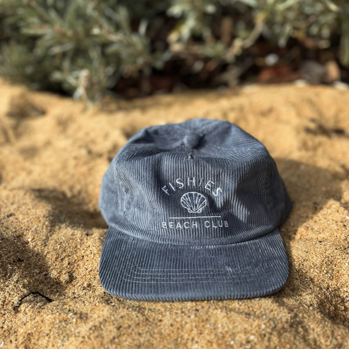 Blue cap with 'Fishers Beach Club' logo kids cord cap on sandy ground with plants in the background