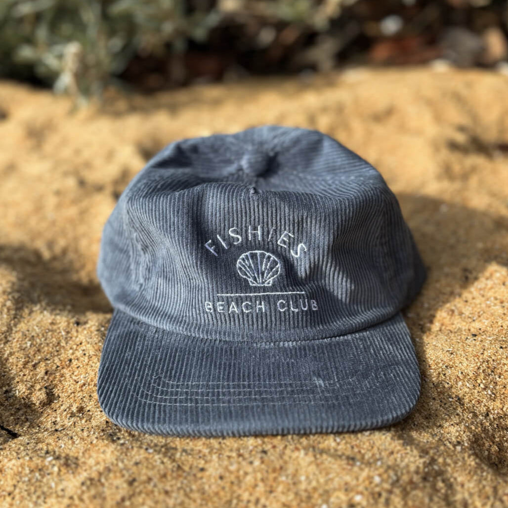 Blue cap with 'Fishers Beach Club' logo kids cord cap on sandy ground with plants in the background