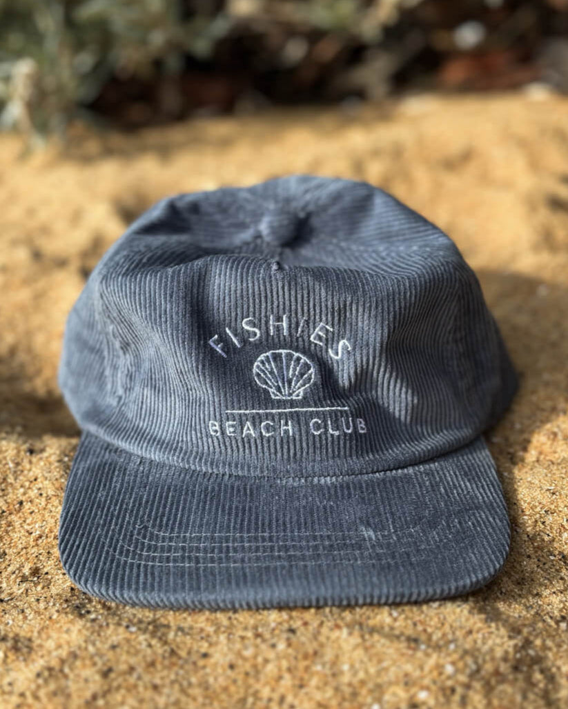 Blue cap with 'Fishers Beach Club' logo kids cord cap on sandy ground with plants in the background