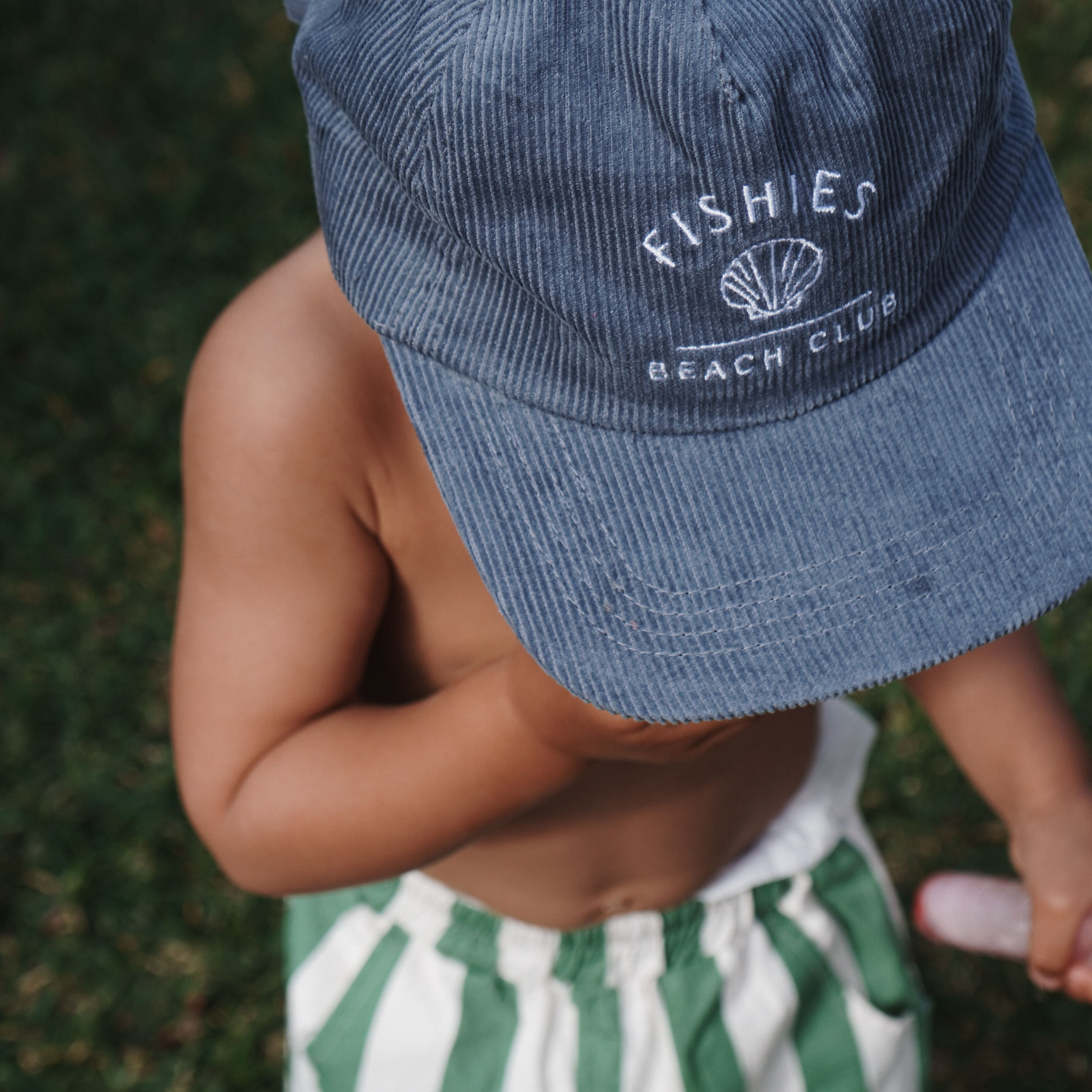 Child wearing a blue cap with 'Fishies Beach Club' logo and green and white striped shorts on grass.