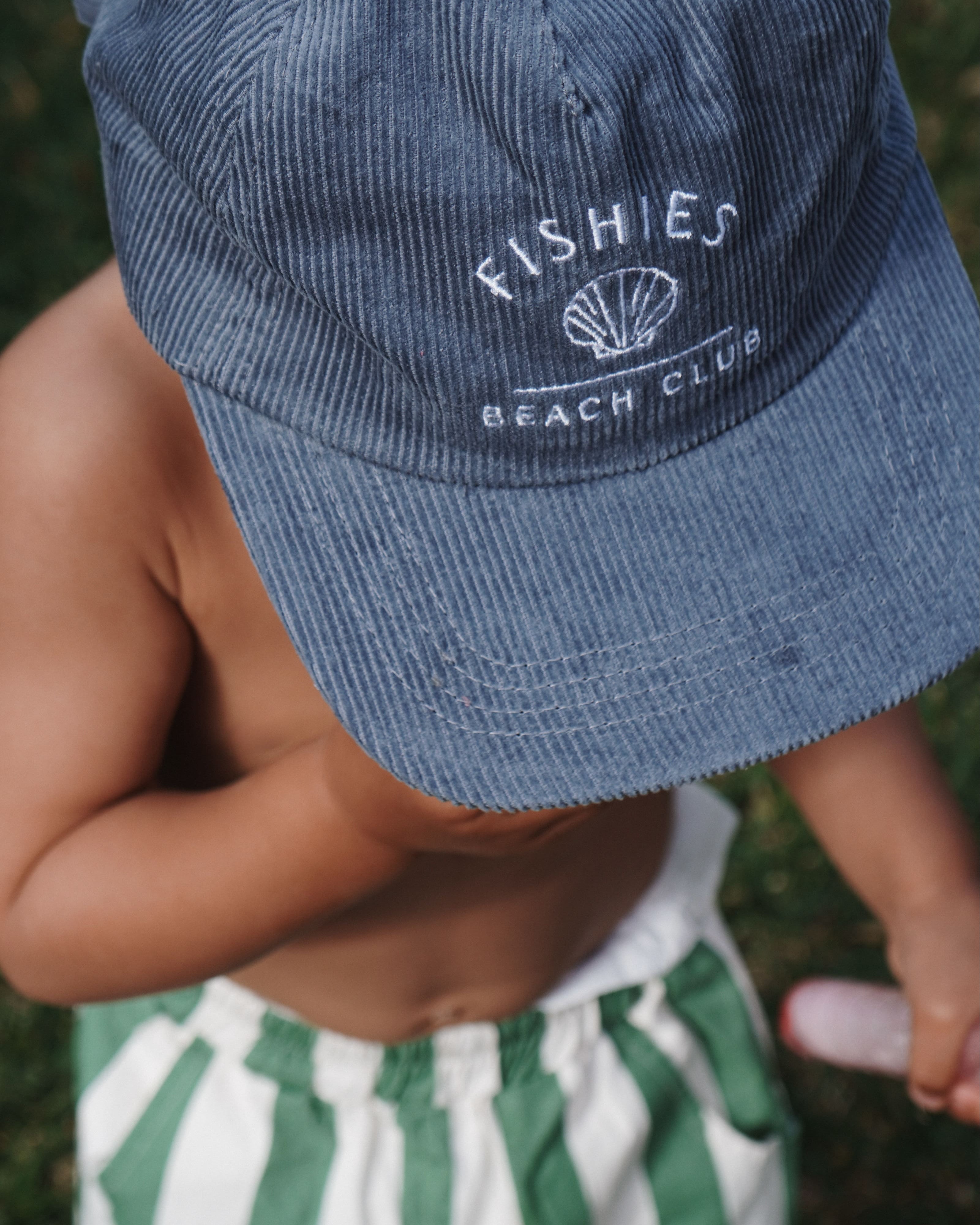 Child wearing a blue cap with 'Fishies Beach Club' logo and green and white striped shorts on grass.