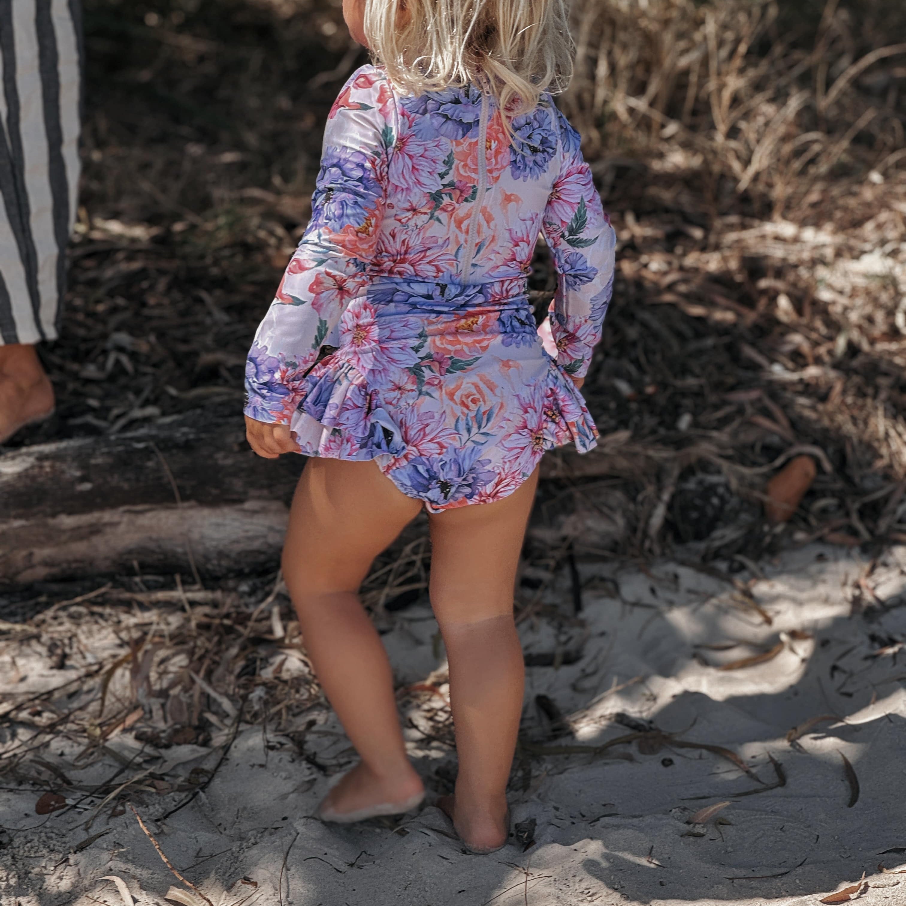 Child in a floral dress standing on a sandy beach with dry grass and twigs wearing a long sleeve upf50 sun safe swimsuit with no toxins and nappy change snapsaround.