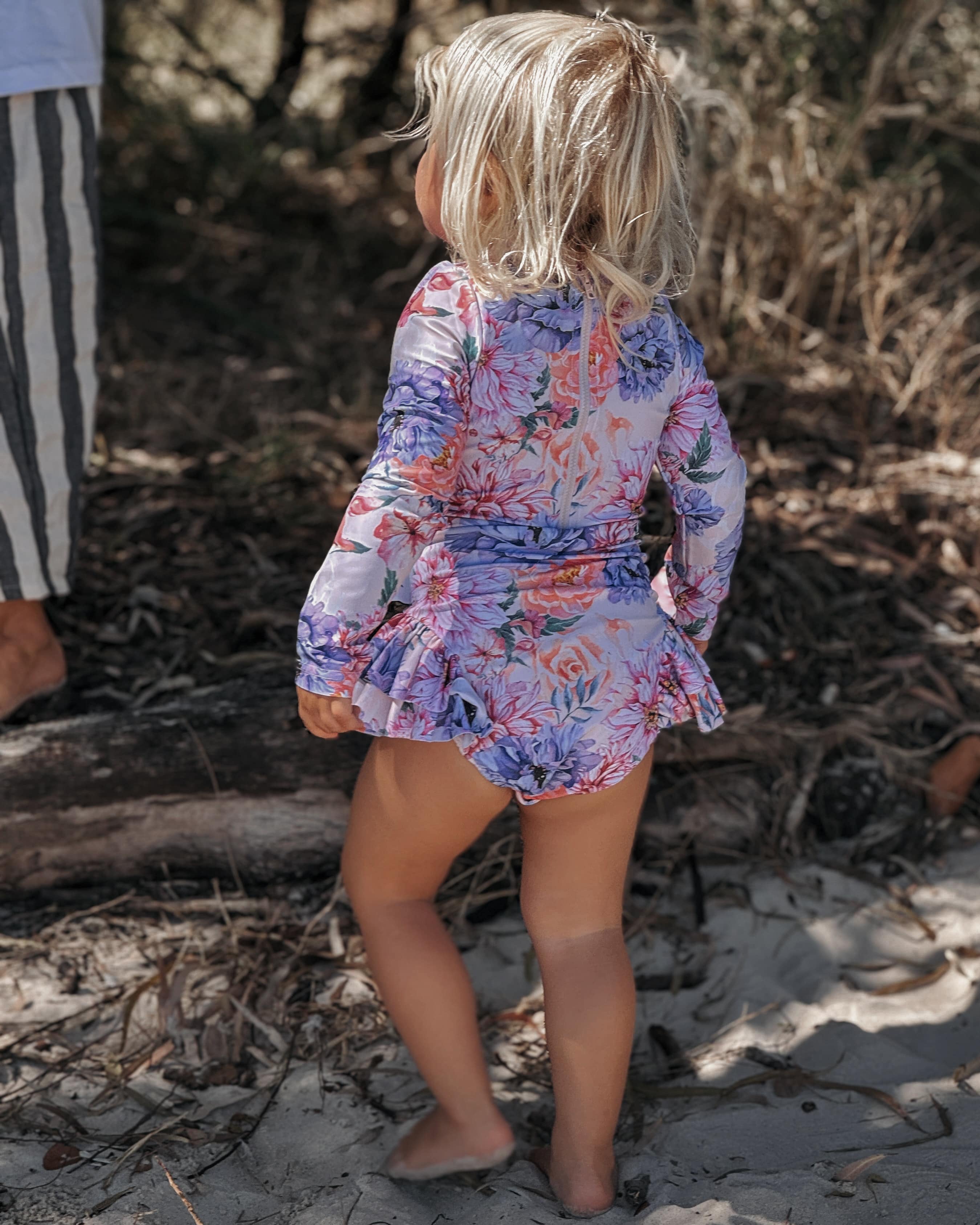 Child in a swimsuit with corth snaps standing on a sandy beach with dry grass and twigs around.