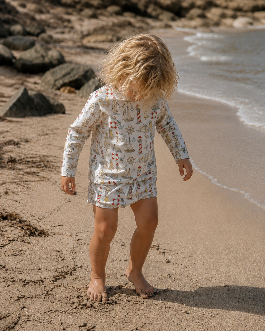 Child in a floral outfit walking on a beach with rocks and water in the background