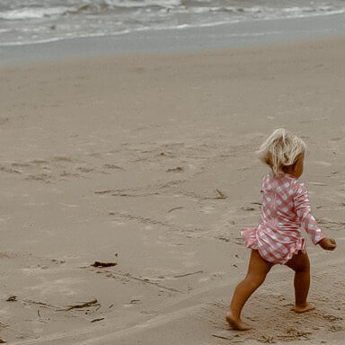 Child in a pink checkered outfit running on a sandy beach with waves in the background