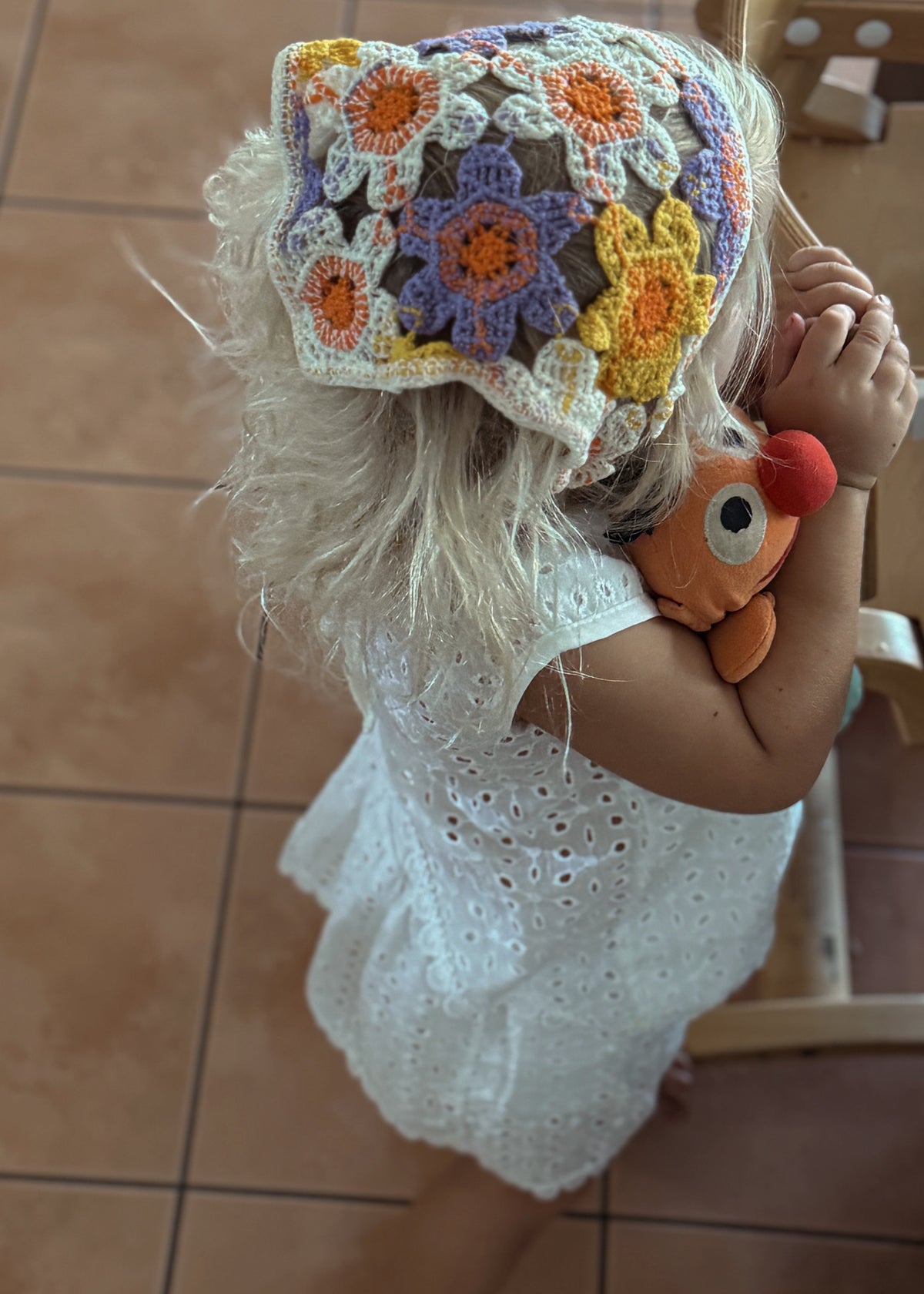 A toddler in a white eyelet dress stands indoors on a tiled floor next to a wooden chair, wearing a colorful crocheted sun hat with orange, yellow, purple, and blue flower motifs.