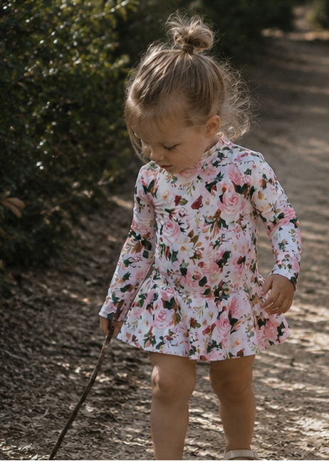 Two young girls wearing matching floral long-sleeve dresses walking barefoot on a dirt path outdoors.