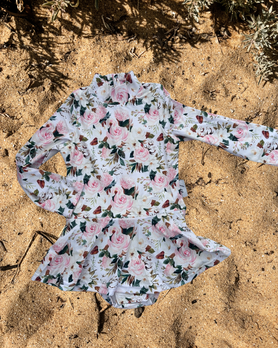 A long-sleeve girls swim suit dress with changing snaps with a pink rose and green leaf floral print, laid flat on sandy ground with some dry grass and shadows visible.