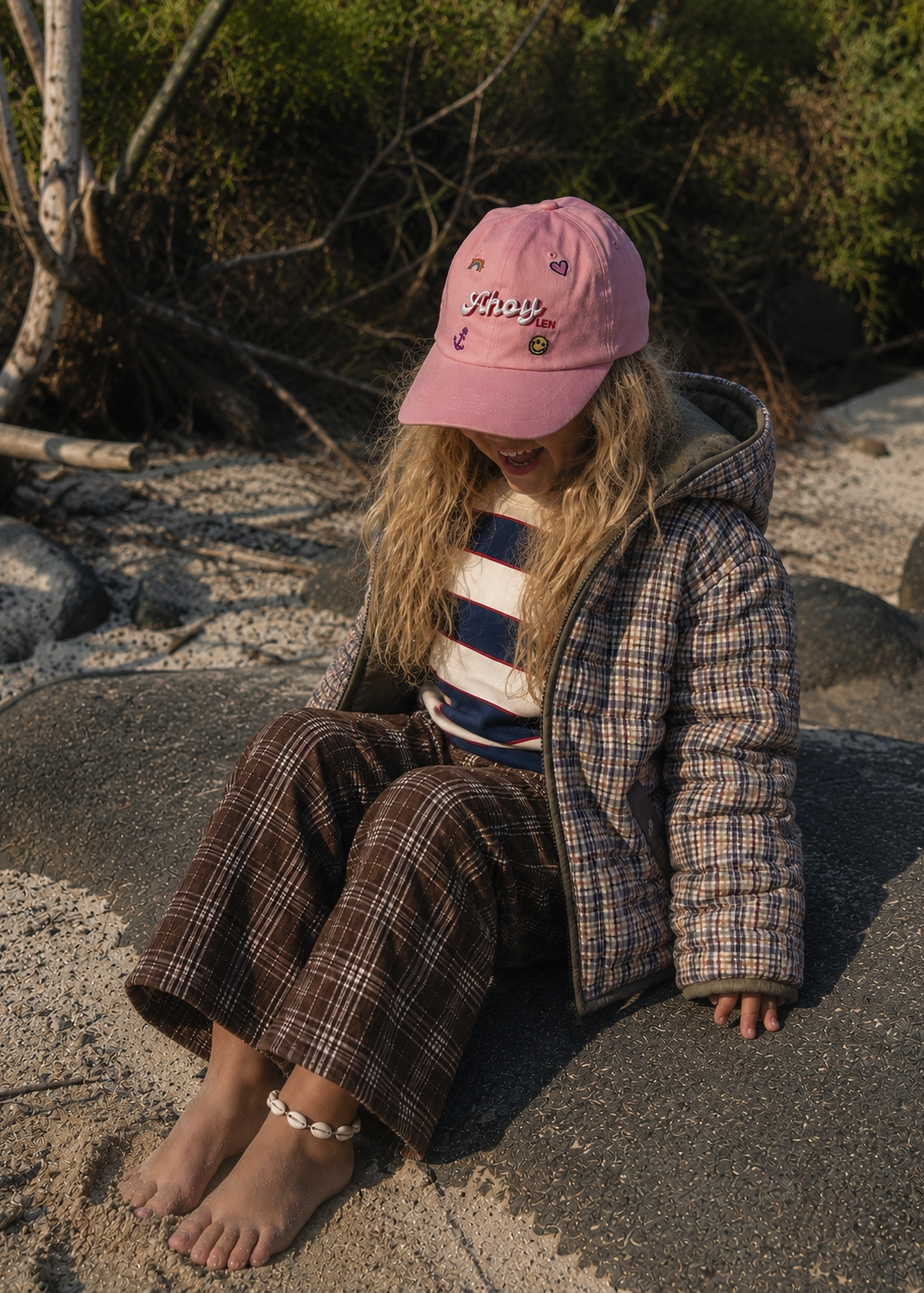 A young girl sits barefoot on a large rock outdoors, wearing a pink baseball cap with 'school' embroidered on it, a striped top, and a brown plaid hooded jacket and matching pants.