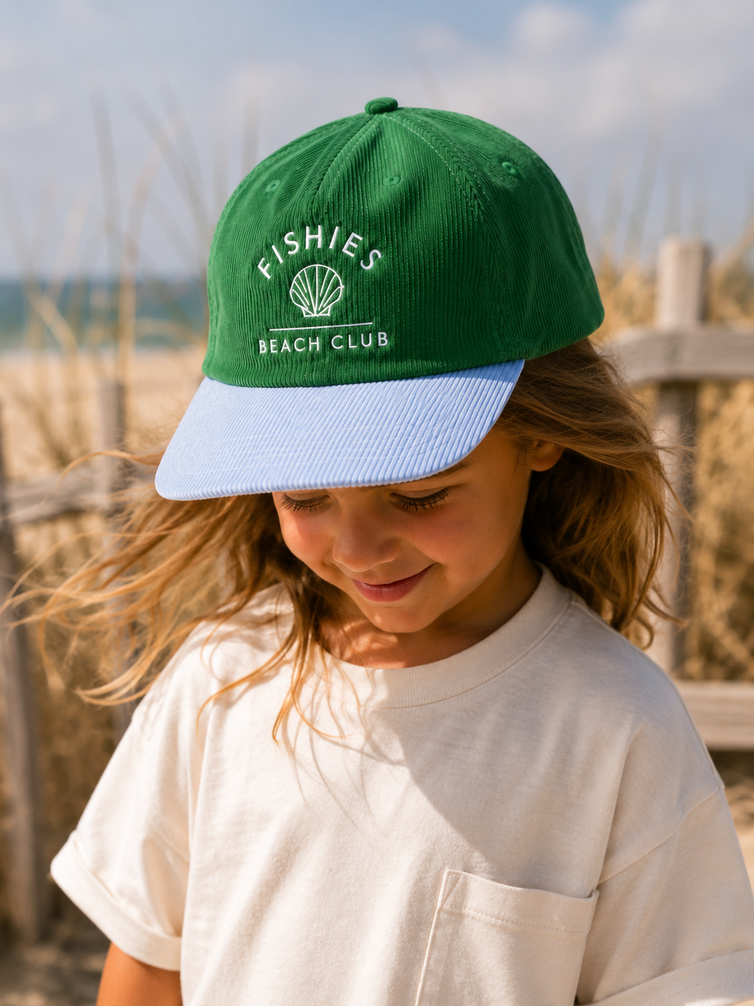 A child wearing a green corduroy baseball cap with a light blue brim. The cap features white embroidered text reading 'FISHIES BEACH CLUB' with a seashell graphic.