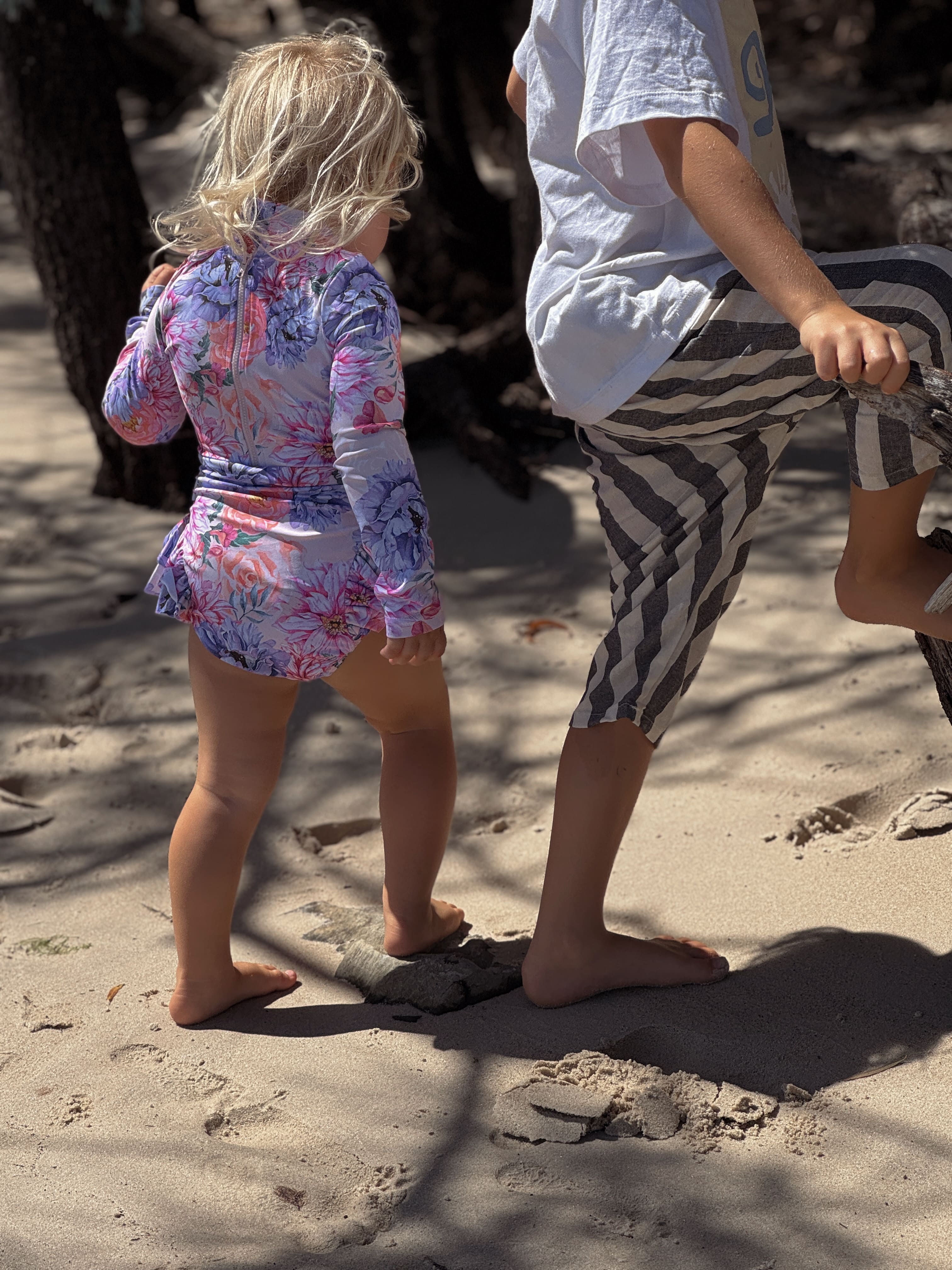 Two children walking on a sandy surface, one in a colorful girls nappy change swimsuit and the other in boys lightweight shorts for summer.