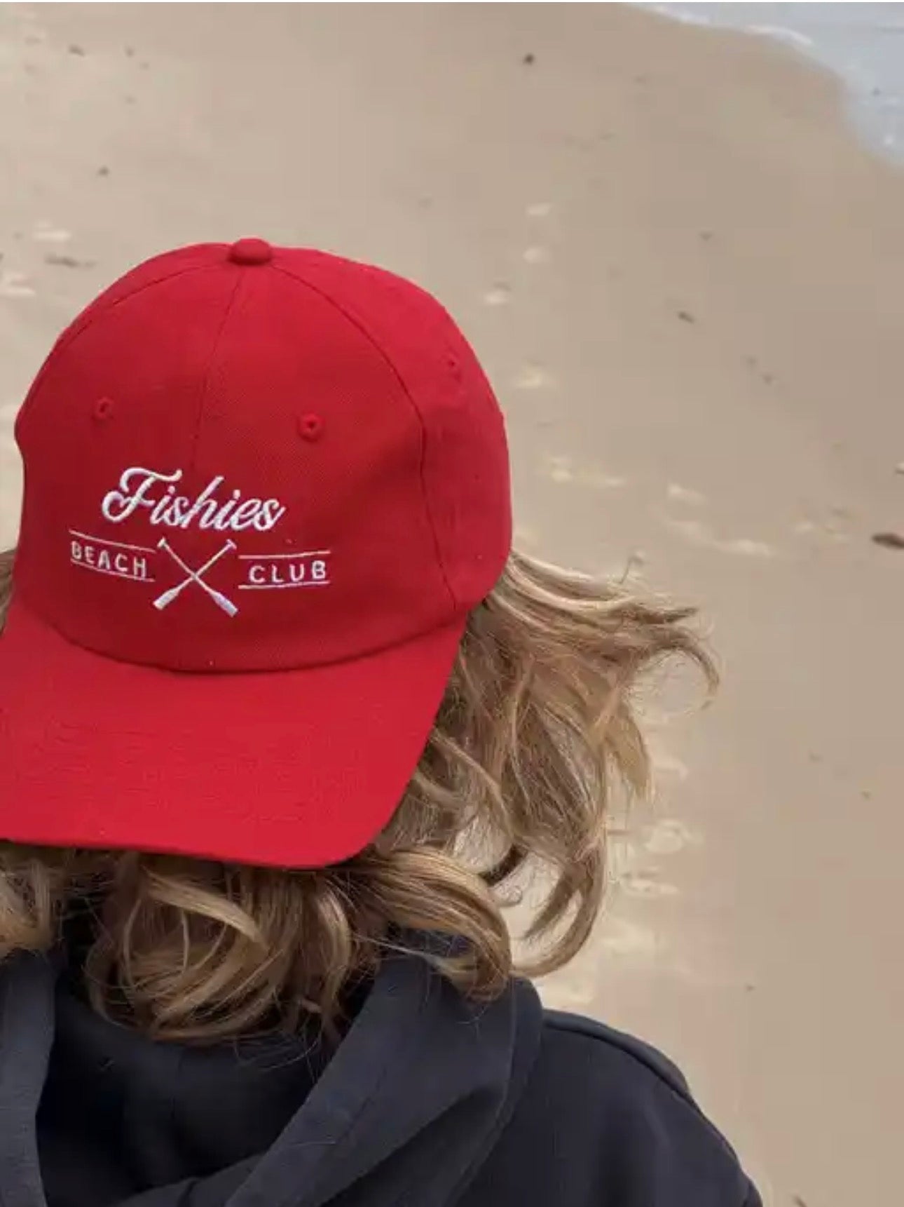 Person wearing a red cap with 'Fishies Beach Club' text on a sandy . cap is a kids cap and bright red background