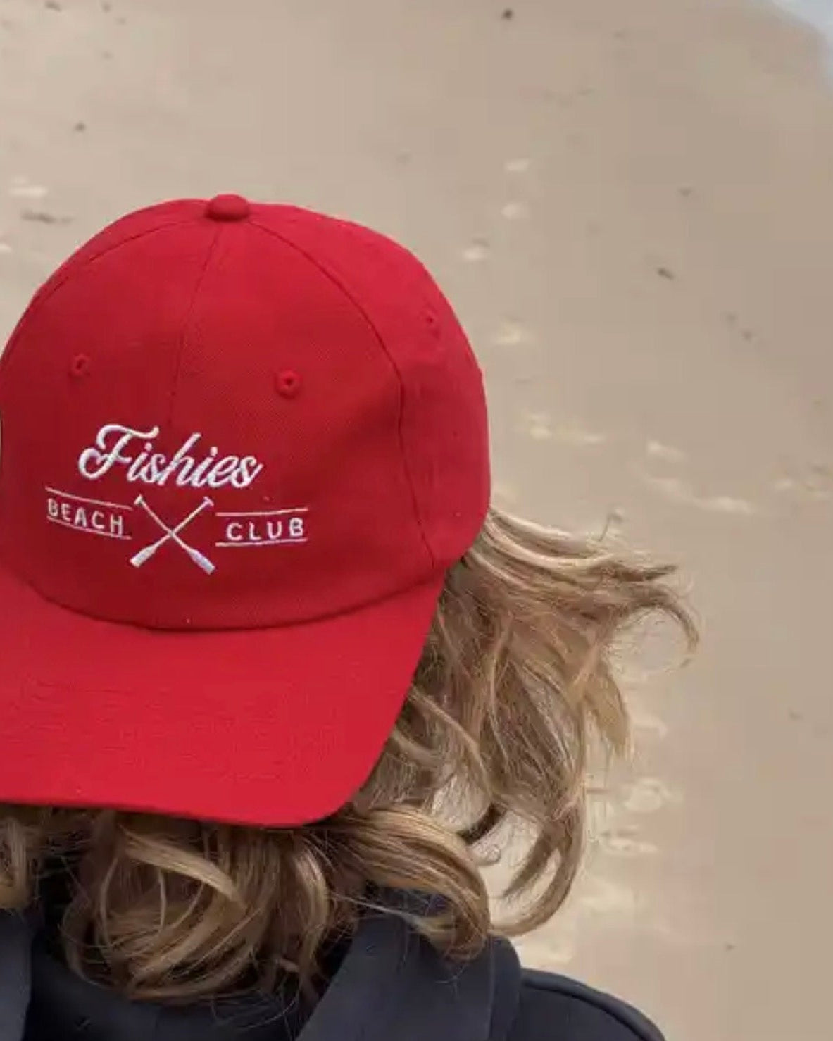 Person wearing a red cap with 'Fishies Beach Club' text on a sandy . cap is a kids cap and bright red background