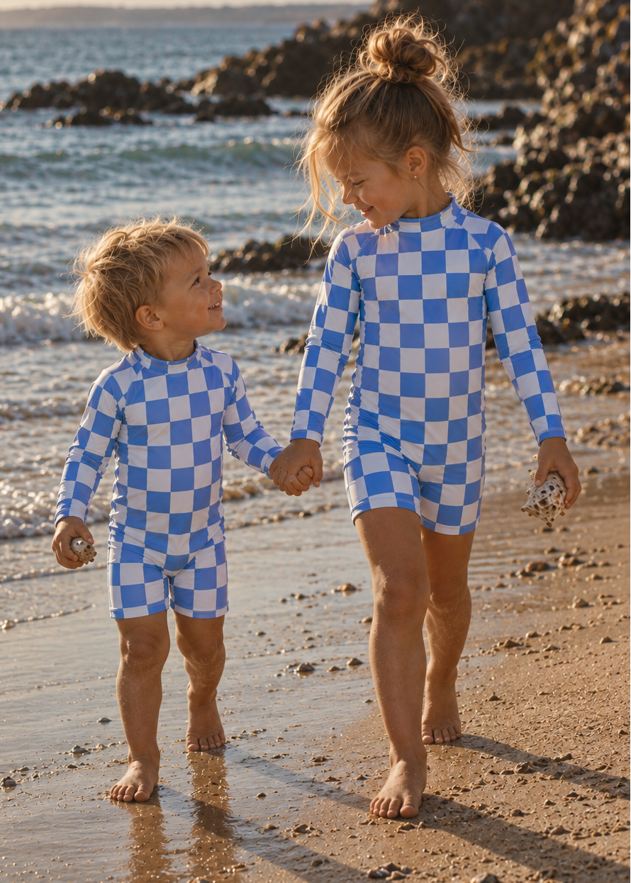 Two children walking hand in hand on a sandy beach wearing matching blue and white checkered long-sleeve swim suits.