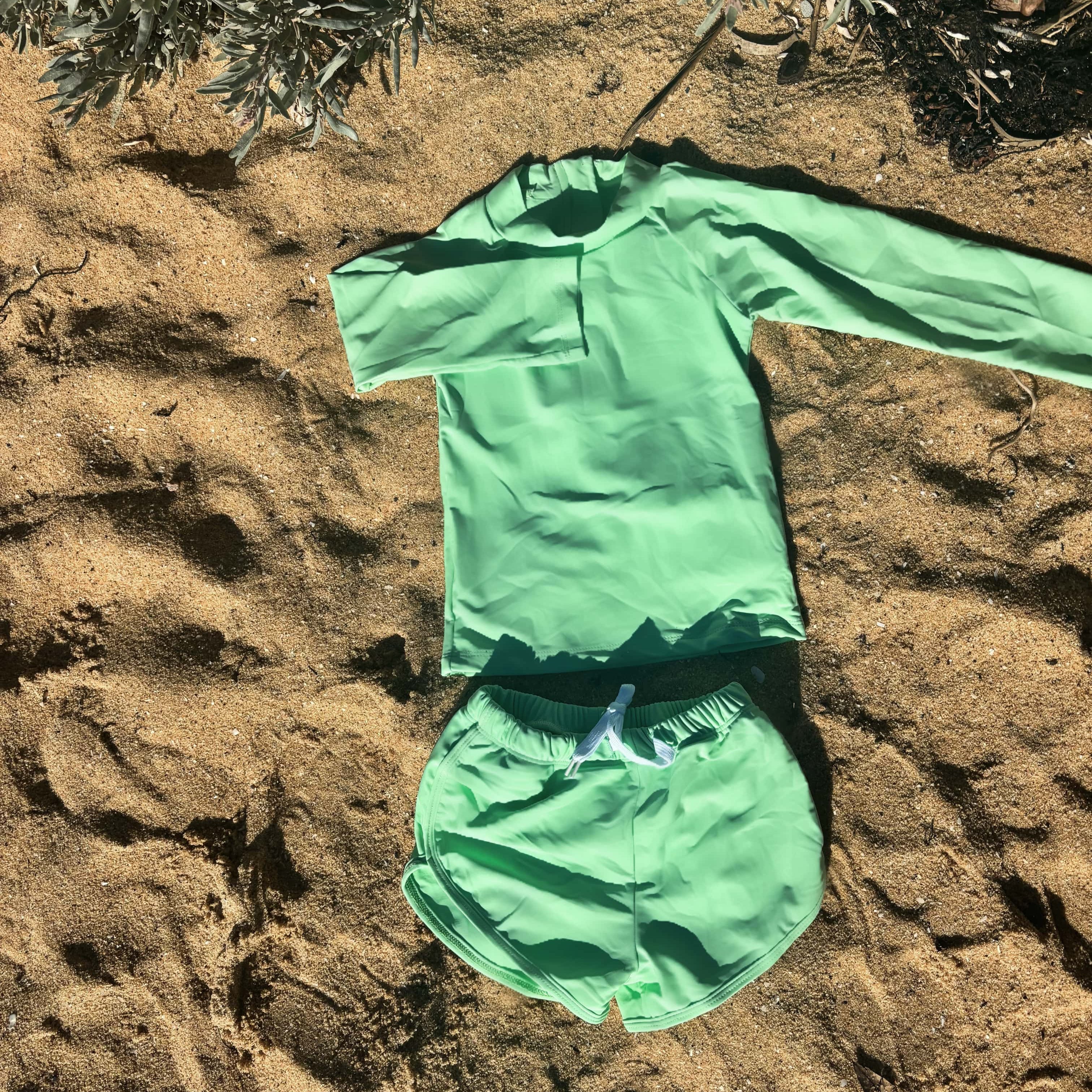 Green bright children's swimsuit set on sandy ground with plants in the background
