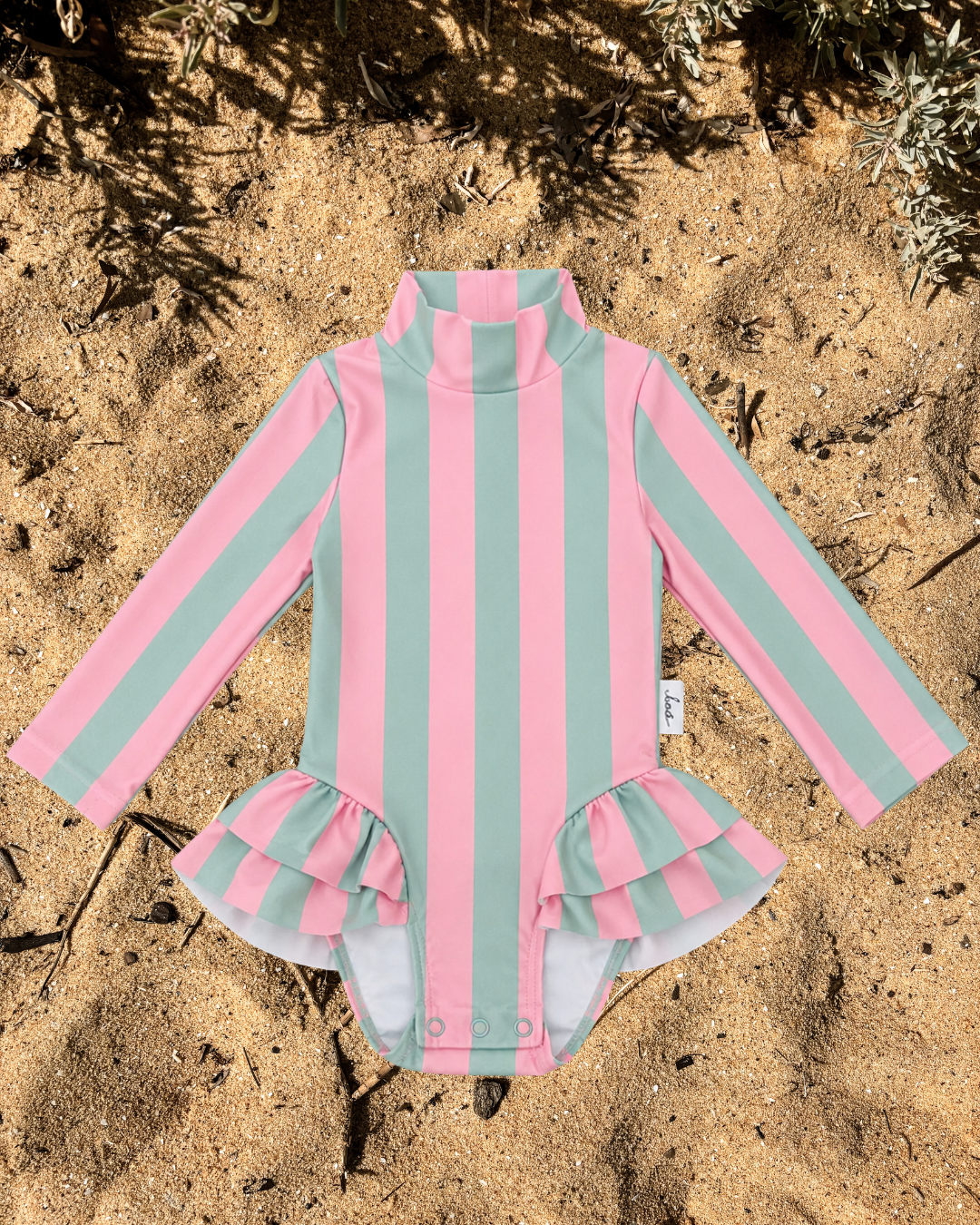 A long-sleeve girls upf50 with vertical pink and mint green stripes, featuring a mock neck and a ruffled leg opening, laid flat on sandy ground with some dry leaves in the background.